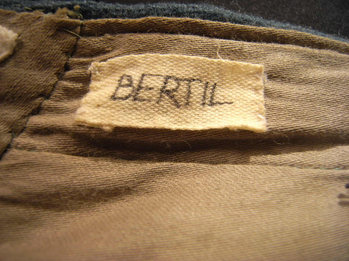 Tunika/kolt av ulltyg, kläde, i duvblå fräg. En bred bård av ett mönstervävt tyg i vit och ljusblå färg längs nederkant, ärmar och bål. Långa ärmar av ett tyg i röd färg. 
