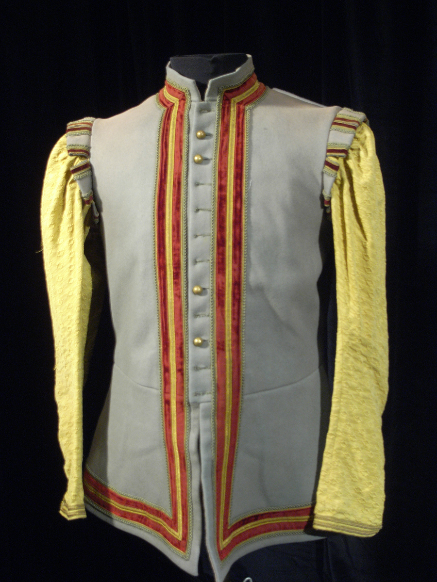 Jacka av kläde i en ljusgrå färg med långa ärmar av mönstervävt tyg i gul färg. Jackan har längre skört, knäppning mitt fram med runda tätt sittande guldfärgade knappar, markerade axelvingar. Dekorband i guldfärg, sammetsband i röd färg samt snoddar i ljusgrå färg.
