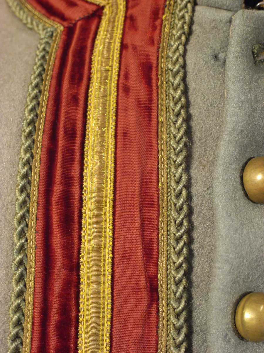 Jacka av kläde i en ljusgrå färg med långa ärmar av mönstervävt tyg i gul färg. Jackan har längre skört, knäppning mitt fram med runda tätt sittande guldfärgade knappar, markerade axelvingar. Dekorband i guldfärg, sammetsband i röd färg samt snoddar i ljusgrå färg.
