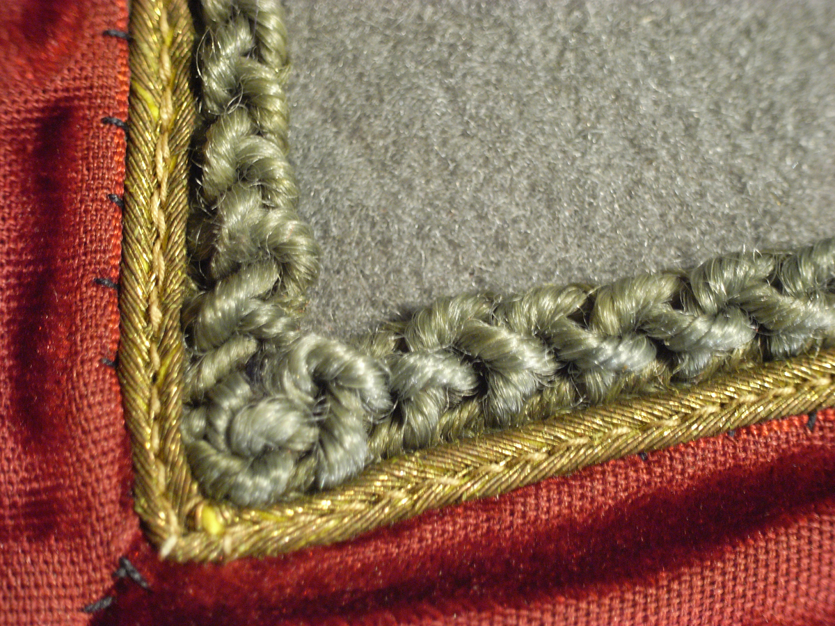 Jacka av kläde i en ljusgrå färg med långa ärmar av mönstervävt tyg i gul färg. Jackan har längre skört, knäppning mitt fram med runda tätt sittande guldfärgade knappar, markerade axelvingar. Dekorband i guldfärg, sammetsband i röd färg samt snoddar i ljusgrå färg.
