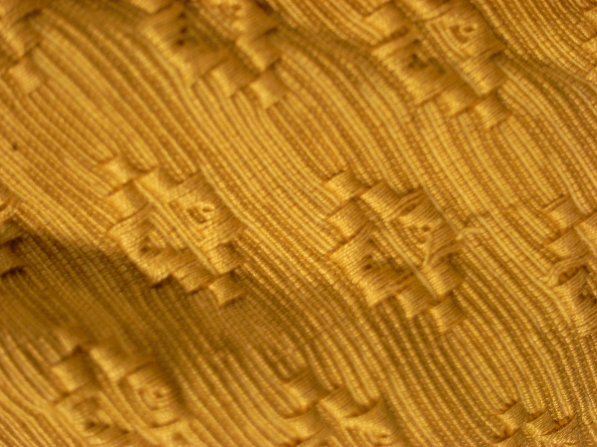 Jacka av kläde i en ljusgrå färg med långa ärmar av mönstervävt tyg i gul färg. Jackan har längre skört, knäppning mitt fram med runda tätt sittande guldfärgade knappar, markerade axelvingar. Dekorband i guldfärg, sammetsband i röd färg samt snoddar i ljusgrå färg.
