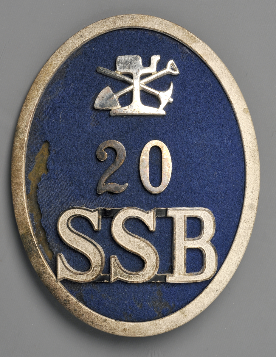 Mössmärke för pälsmössa, ovalt med ram av nysilver och Ljusblå klädes-botten. Med banavdelningens emblem, Siffran 20 samt SSB i silver. För banpersonal.
SSB
