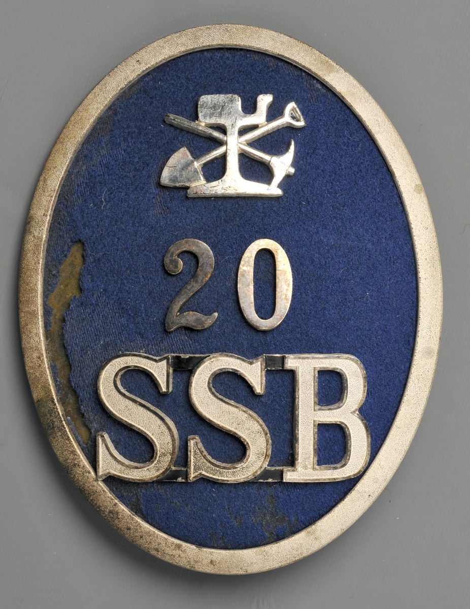 Mössmärke för pälsmössa, ovalt med ram av nysilver och Ljusblå klädes-botten. Med banavdelningens emblem, Siffran 20 samt SSB i silver. För banpersonal.
SSB