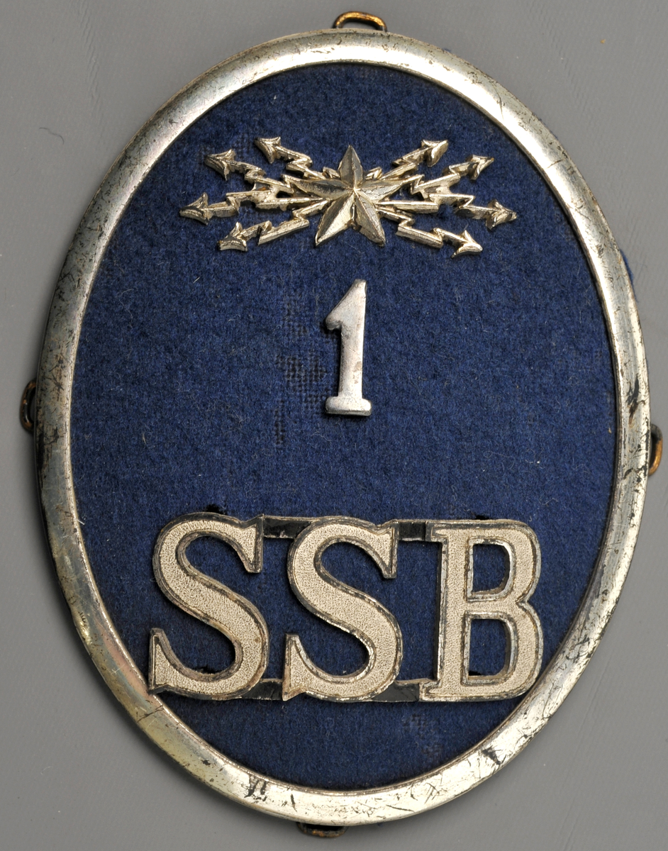 Mössmärke för pälsmössa, ovalt med ram av nysilver och ljusblå klädesbotten. Med ledningsavdelningens emblem (stjärna med blixtar), siffran 1 samt SSB i silver. För ledningspersonal.
SSB