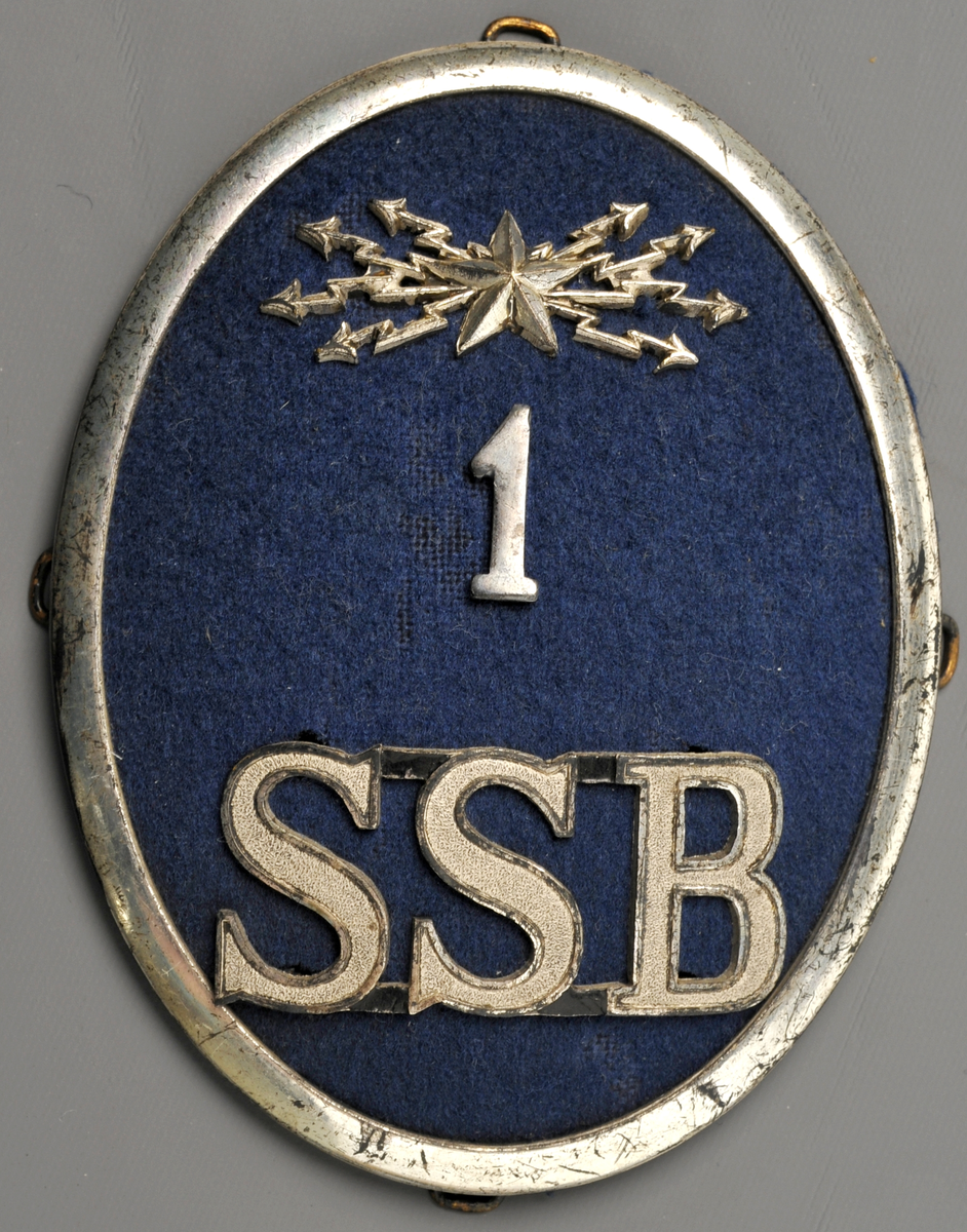 Mössmärke för pälsmössa, ovalt med ram av nysilver och ljusblå klädesbotten. Med ledningsavdelningens emblem (stjärna med blixtar), siffran 1 samt SSB i silver. För ledningspersonal.
SSB