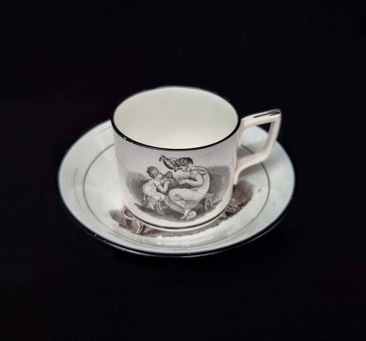 Kaffekopp med fat i pearlware.

Tillverkad i fajans med vit glasyr som har en svag blå ton, typisk för pearlware – en keramisk teknik utvecklad i England av Josiah Wedgwood omkring år 1779. Den blå tonen i glasyren uppnås genom tillsats av kobolt.

Dekoren är utförd med transfertryck och föreställer kvinnor med barn. Motivet är nyklassicistiskt och var vanligt under den tidiga empiren, omkring 1800–1810.

Det är inspirerat av den irländske konstnären Adam Buck (1759–1833), känd för sina idealiserade familjescener.

Troligen tillverkad i Staffordshire, England.