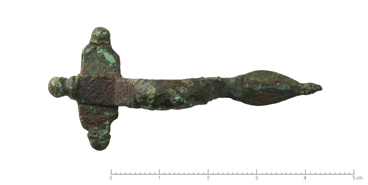 "2251 a. Liden korsformet bøilenaal af bronce, hvis nedredel er udvidet til en dyrehoved-lignende plade uden antydning til øine; den er kun 5,5 cm lang." Liten, korsformet spenne av bronse med små knopper, flate på baksiden støpt i et med spennen. Hodeplaten måler 25 x 8 mm. Den har et opphøyet midtfelt i forlengelsen av bøylen. Bøylen er jevnbred, flat på baksiden og konveks på fremsiden, stbr bøyle: 6 mm. Foten er flat, nærmest formet som et blad, med midtrille. Foten ender i en profilert, liten spiss. I overgangen til plate og fot er bøylen dekorert med tversgående riller. Spennen er en del korrodert, den mørkrøde fargen er kobberkorrosjon.