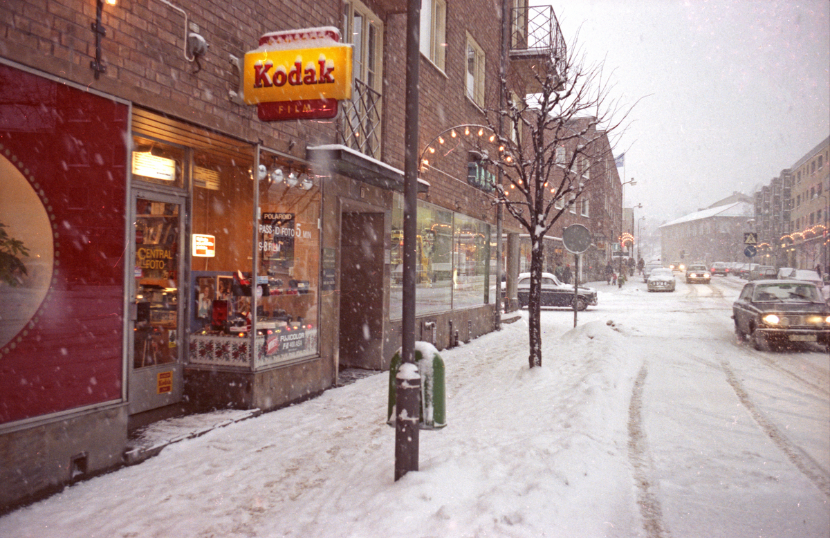 Centralgatan 7 vid Centralfoto (Tidigare Atelier Ekegren), kvarteret Postmästaren. Ovanför skyltfönstren sitter en skylt med texten "Kodak". På Centralgatan ligger nyfallen snö och på husfasaden sitter juldekoration i form av bågar med lysande lampor. Vinter. 