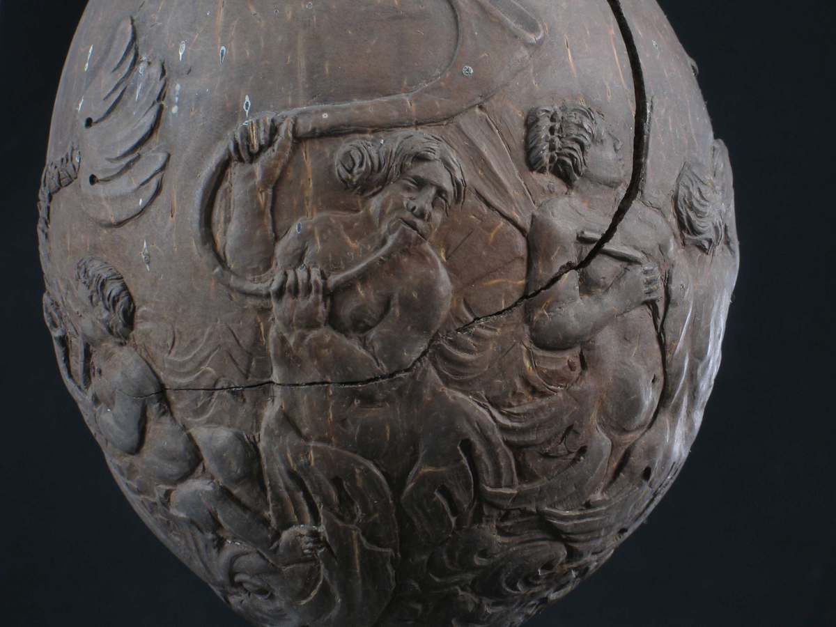 Foten har fire ornamentfelt i relieff, to av dem med blomsterdekor, de to andre med blad og overflødighetshorn. Pokalen bæres av en Neptunfigur utskåret i elfenben, med løftet venstrearm (denne har hatt scepter) og delfin mellom benene. På Neptuns hode en pokal av en kokosnøtt med toppen avskåret som lokk, og med beninnfatning.  Pokalen har relieffutsmykning med nereider og tritoner, som blåser i en koncha (basunlignende instrument). Lokket har fire relieffmotiver med nøtter og to med blomster.  Lokkgrep av ben, balusterformet, med sittende apekatt i metall på toppen.