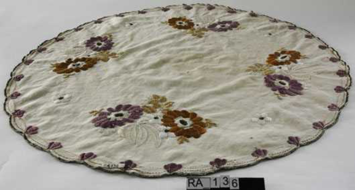 Duk, ecrufarget, sirkelformet med tunger. Broderi sort omkretskant, fire rosebuketter i oransj og fiolett, rosenknopper langs ytterkant.