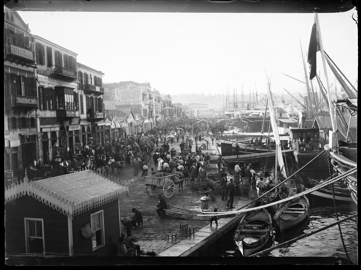 "Brygge, Smyrna." (Bildetekst fra album) Stor folkemengde på en havn. Hester og kjerrer, varer og båter.