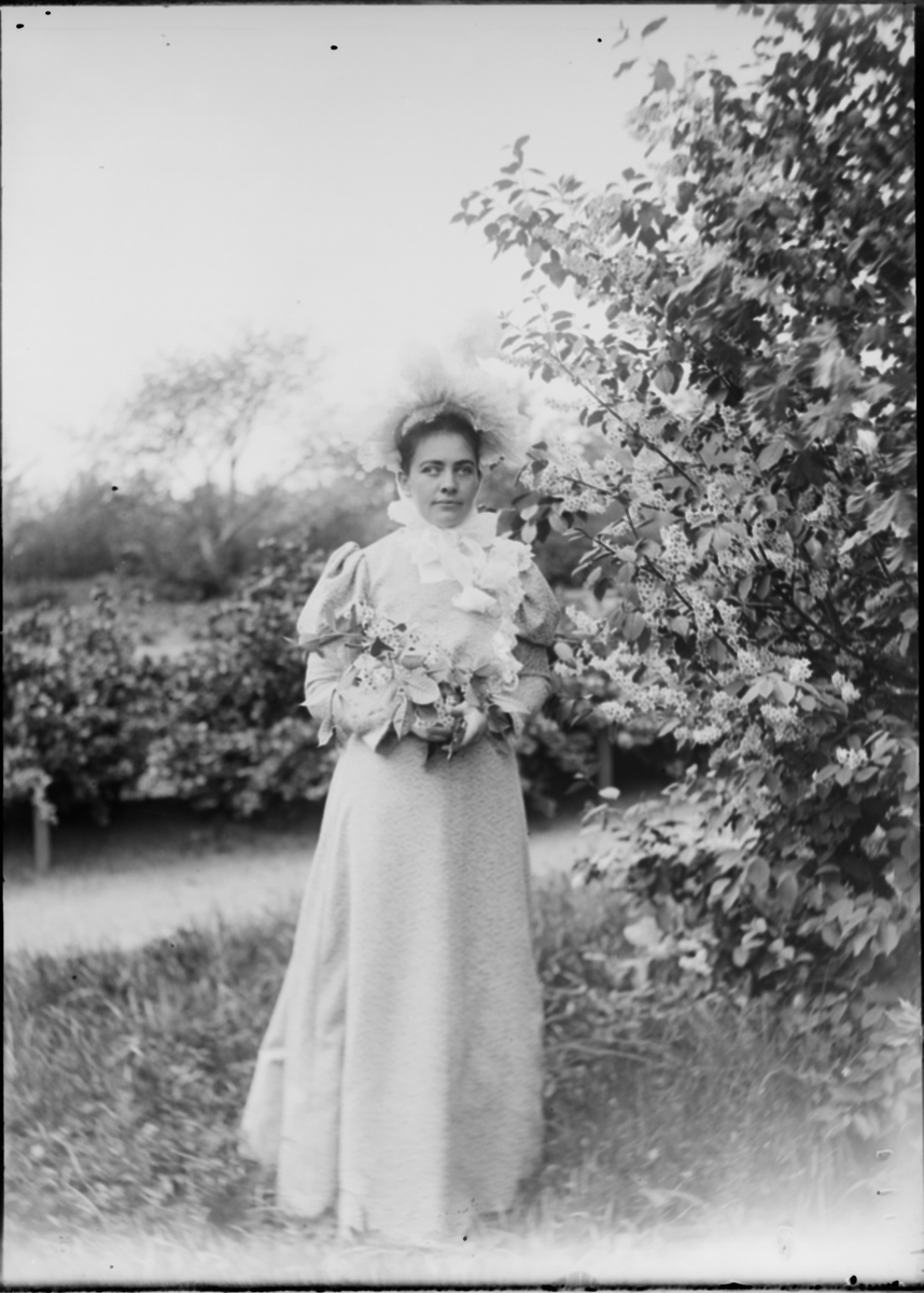 "Julie Mathiesen." (Bildetekst fra album) En ung dame i en hage med blomster i hendene.