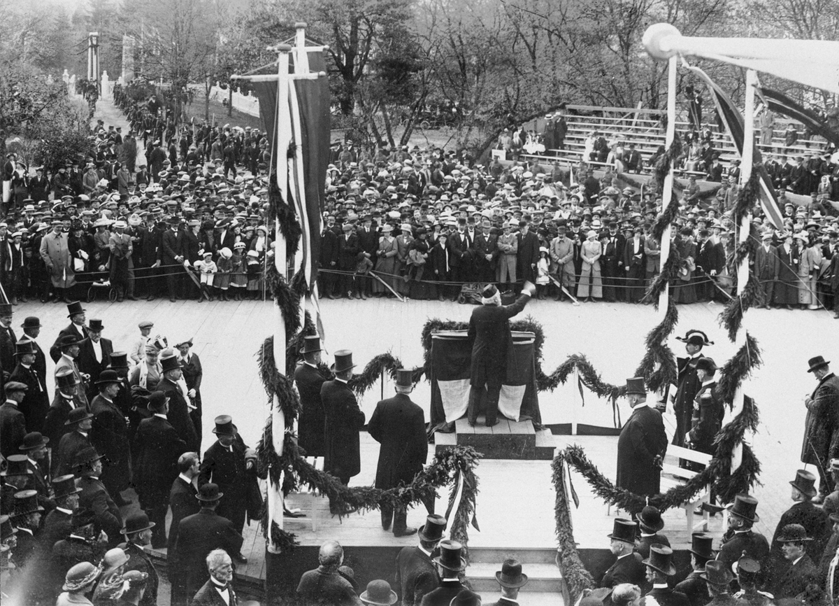 Den 17. mai ble det holdt flere taler foran Eidsvollbygningen. Talen for dagen ble holdt av stortingspresident Jørgen Løvland, og det er trolig han som er taleren på dette bildet. Kong Haakon står til høyre for taleren med en hatt som skiller seg klart fra flosshatter og skalker. Bildet er tatt fra Eidsvollbygningen. I bakgrunnen porten og oppkjørselen til bygningen. Helt til høyre en tribune som var bygd for anledningen.
