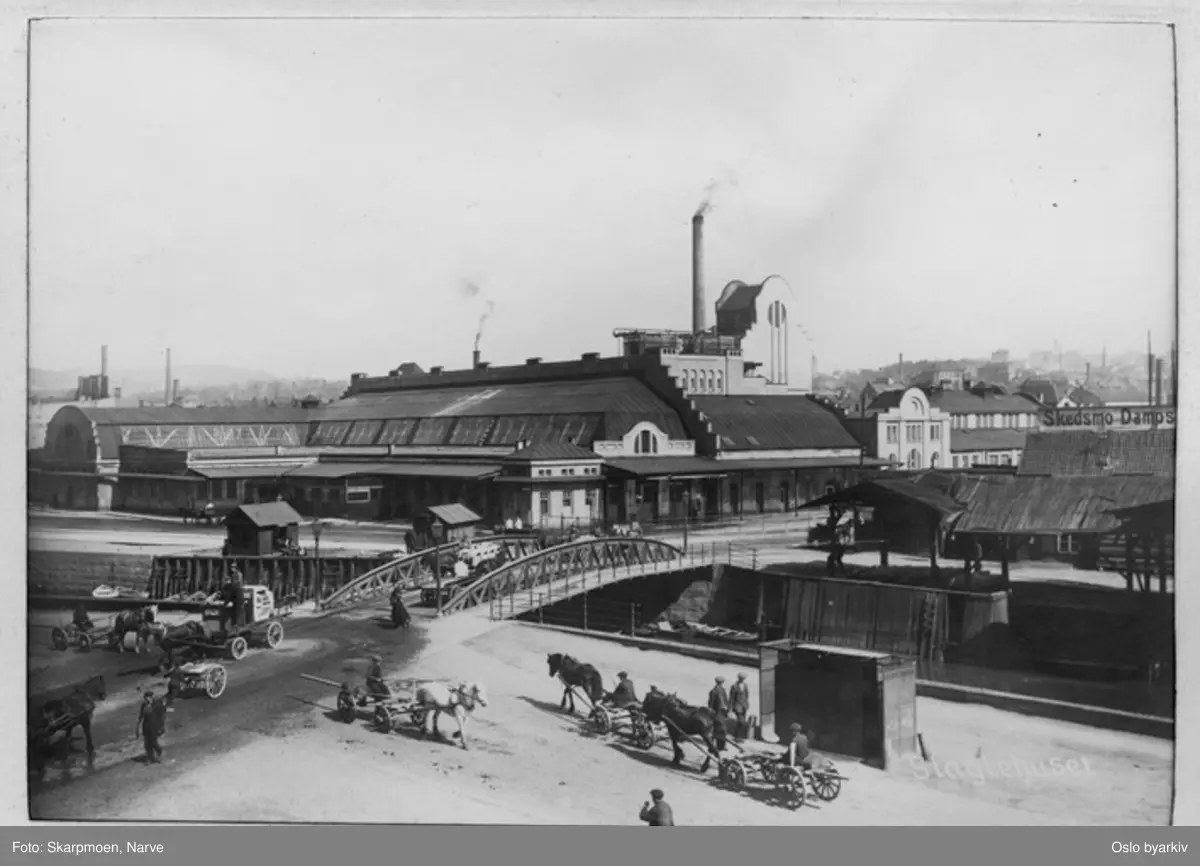 Schweigårds bru over Akerselva til slaktehuset på Grønland, mange hester med vogn, menn i gatebildet, røyk fra pipene på slakteriet.