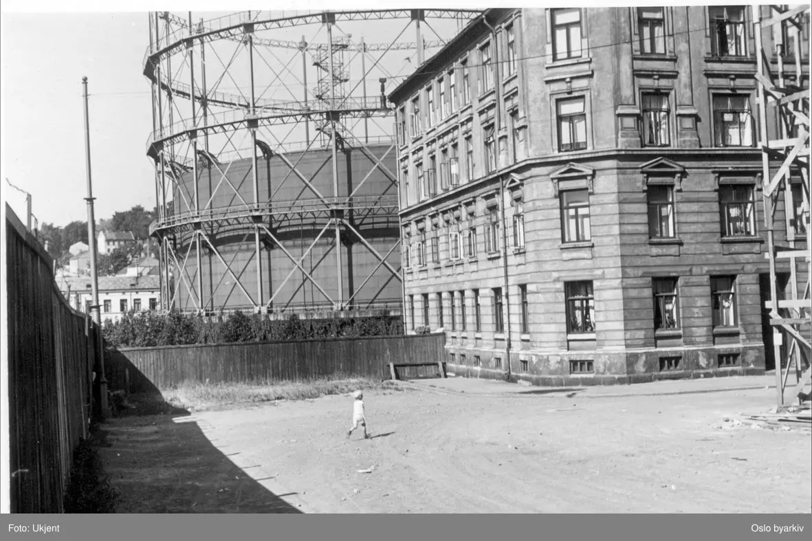 Grüner gate langs og til plankegjerdet. Marselis' gate inn fra høyre med bygården (leiegårdskompleks) Marselis' gate 26 (gården i elva) et trefløyet anlegg i fire etasjer og med to underetasjer mot og helt inntil Akerselva. I dag parkbelte. Oslo Lysverkers store gassklokke ruvende bak.