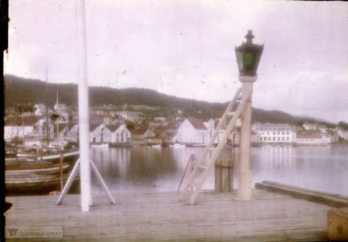 Fra østenden av den gamle Storkaia, som var bygd i tre. Parafinlykta ble tent hver kveld og tjente både til å lyse opp kaia og som kjennetegn for båtene..Bildet er trolig tatt rundt 1910. .(Kopiert fra Lumiere-Autokrome plate, diapositivmateriale i bruk fra 1907.)