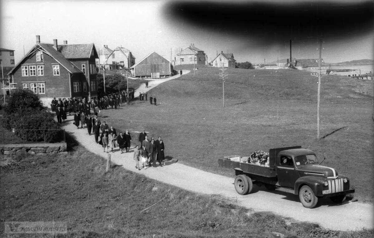 Fra Ingebrikt Huse sin begravelse..Jon Opstad sin lastebil kjører kisten mens gravfølget går fra Steinshamn til kirken..I bakgrunnen f.v ser vi Sæterhaug garden (Olaus S), I.P Huse sitt internat og messe, Olav Skotheim, I.P Huse sin løe, Stornes, Thomas Sandø..Ford V8 1946-47. Det lille merket på døra er sannsynligvis et lønneblad, Canadas nasjonalsymbol, og viser at bilen er fra et parti kanadiskbygde Forder som ble tatt inn like etter krigen..(O.Skotheim dreiv landhandel i kjerraren i garden tidligere I.P Huse)