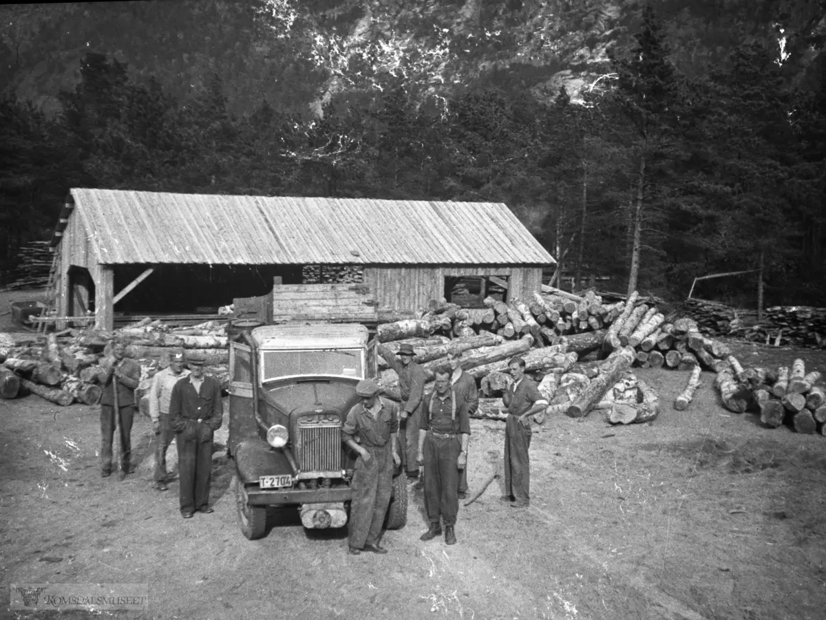 Saga i bakgrunnen bygde Håkon E Utigard og Henry Finnset i 1944..Bil med reg nr T-2704..1933-34 (Chevrolet?) Rugby lastebil med frontmontert Imbert generatoraggregat, for kjøring med knott. .Det kan tyde på at bildet er tatt under eller umiddelbart etter krigen. Tror det var slutt på knottkjøring i løpet av 1946. .I Hvem eier bilen 1948 står T-2704 på Chevrolet lastebil til Ingvar Moen, Elnesvågen..."I Hvem eier bilen 1939 står T-2704 på Eiro Auto, Neste.som Chevrolet lastebil. Årsmodellen må i tilfelle være 1934-35. 1933-modellen hadde i hovedsak samme utseende som den hadde hatt siden 1929. Våren 1935 var regnr T-2704 ennå ikke tatt i bruk, det høyeste i denne Molde-serien var i Norges Bilbok 1935 T-2670. Rugby har ingenting med Chevrolet å gjøre, Rugby lastebiler ble produsert av Durant-konsernet 1928 til konkursen i 1932 og så eldre ut enn denne".