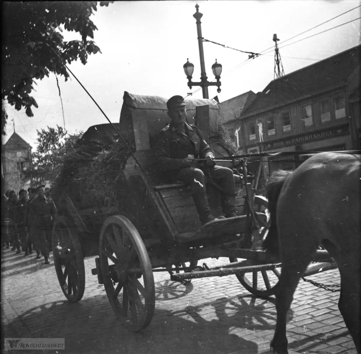 Hest og kjerre er i Kongens gate. .Etter 8. mai 1945? Kusk med tysk uniform? Soldater bak vogn i engelsk uniform?.Husene til høyre er i kvartalet mellom Torvet og Prinsens gate. Bok- og papirhandelen til høyre, er Globus-Forlaget A/S i Kongens gate 11-13. I bakgrunnen, Vår Frue kirke.."F.Bruns Bokhandel ligger i bygningen som skimtes bak trærne bak den antatt engelske soldatene. .Bildet viser Globusforlaget bok- og papirhandel- helt til høyre ser vi litt av fotograf Schrøders gård"..Vår frue kirke ses til venstre.