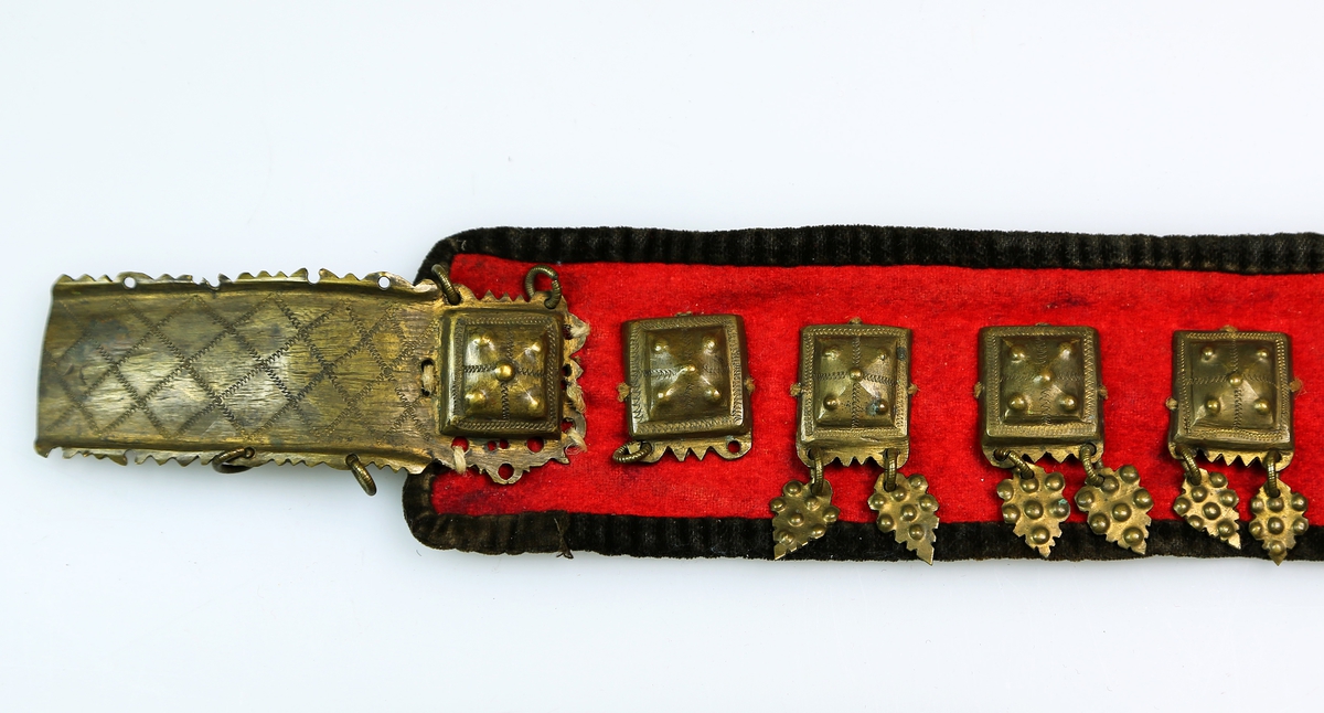 Stølebelte av rødt ullstoff kantet med mørkt ullstoff og med striestoff på innsida. Det er små beltestøler langs hele beltet, som hver har to lauv. Beltespenna har enkel gravert dekor. 