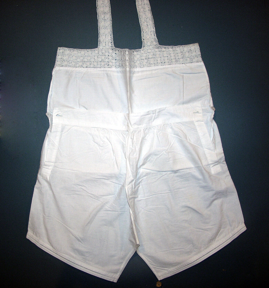 Combination, kvinne. N1b
Ett for-og ett bakstykke, rett oventil, påsydd 6 cm bred heklet kant rundt åpning. Ikke ermer, 3, 5 cm bred heklede skulderstropper. Buksa søm midt foran, 14 l splitt i sidene, kneppet med stofftrukken knapp, søm midt bak, bakstykket til å kneppe ned, 4, 5 cm bred linning, flere små rynkinger på hver side av midtsøm bak. Hullfaldsøm rundt låråpning. Livstykket sprukket i siden fra linning til heklekant.