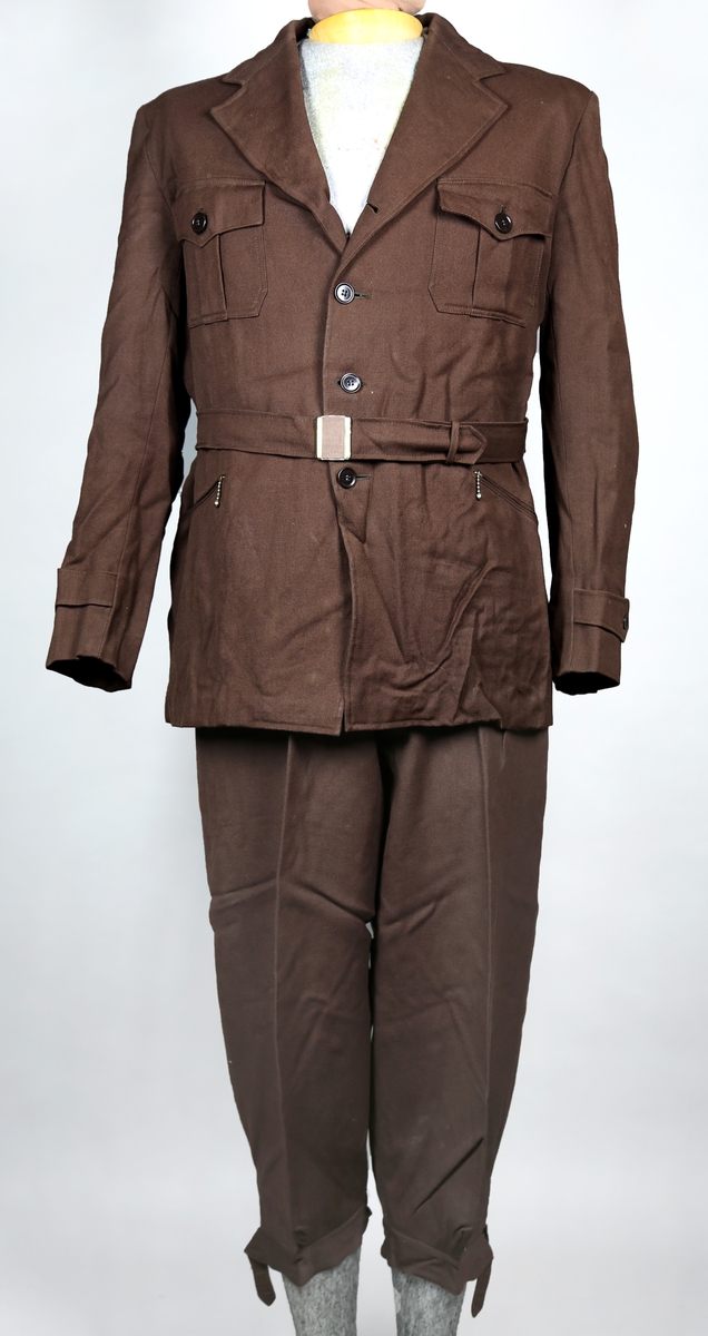 Dress bestående av jakke og bukse. Jakka er enkelt spennet og kneppes med tre knapper, og i tilleggg er det et belte rundt livet. Buksa er kort, - knekort. Jakka har fire lommer utenpå, hvorav de to nedre kan lukkes med glidlås og de to øvre lukkes med knapper. Den er fôret med silke. Buksa har både plastknapper og metallhekte. Nede kan buksa strammes med stropper rundt leggene. 