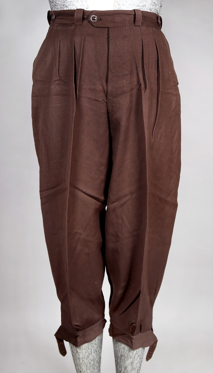 Dress bestående av jakke og bukse. Jakka er enkelt spennet og kneppes med tre knapper, og i tilleggg er det et belte rundt livet. Buksa er kort, - knekort. Jakka har fire lommer utenpå, hvorav de to nedre kan lukkes med glidlås og de to øvre lukkes med knapper. Den er fôret med silke. Buksa har både plastknapper og metallhekte. Nede kan buksa strammes med stropper rundt leggene. 