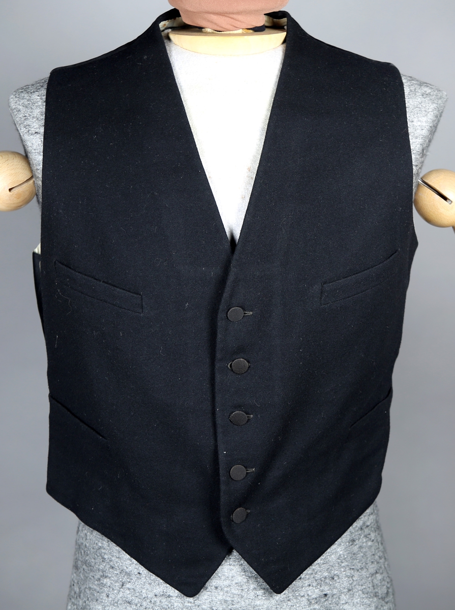 Svart dress med vest og lang jakke. Jakka er dobbeltspennet og kneppes med fire knapper, som er trukket. Jakka har splitt bak. Den har silkefôr, bortsett fra i ermene. Buksa har både plastknapper og metallhekte, og tre lommer. 