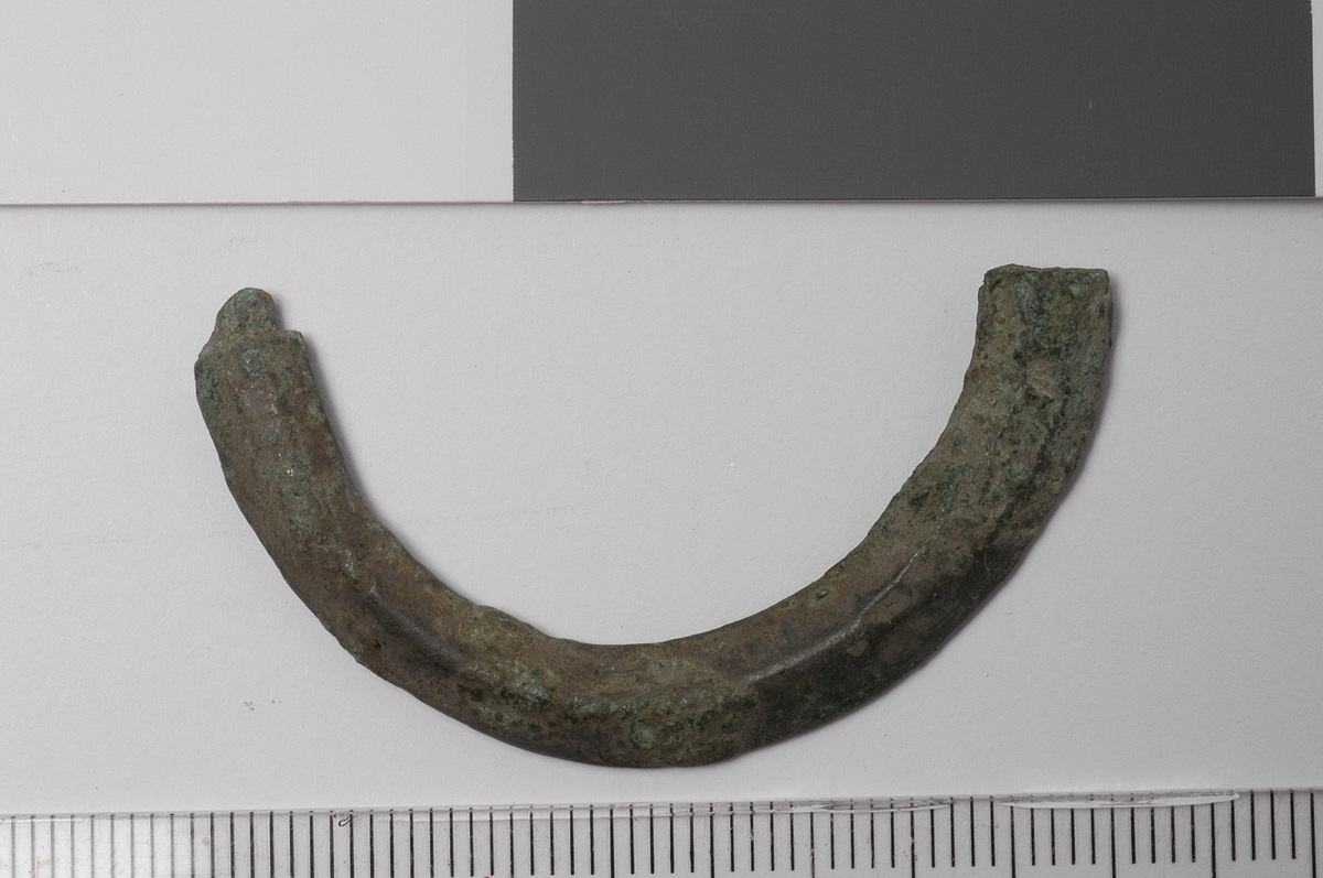 Halvsirkelformet gjenstand med bruddflate i begge ender. Gjenstandens tverrsnitt er trekantet med flat underside og en langsgående rygg midt på oversiden. Dette er en del av en spenne.