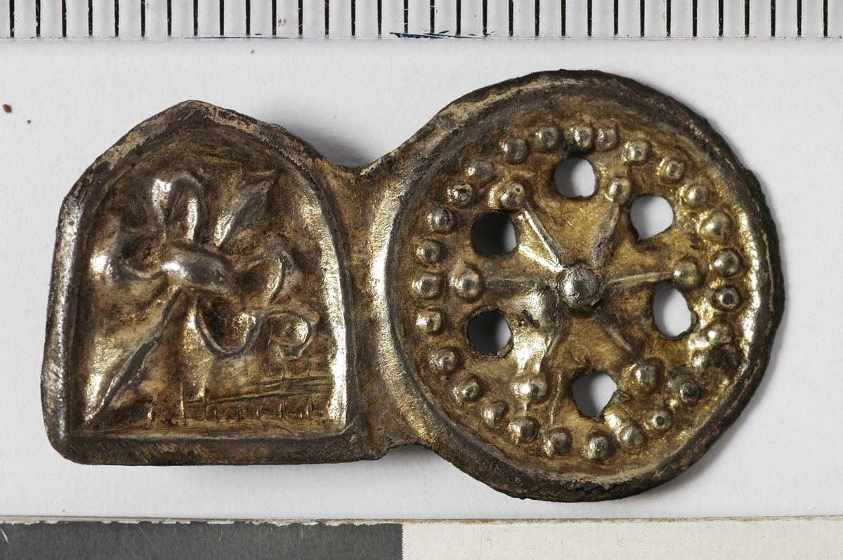 MÅL: L. 27 MM  H. 16 MM, SATT SAMMEN AV ET HJUL OG ET SKJOLD. SKJOLDET ER DEKORERT MED EN FRANSK LILJE, DREVET UT FRA BAKSIDEN. PÅ BAKSIDEN SITTER EN OMBØYD HEKTE. KROK.