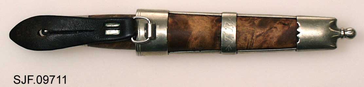 Tollekniv med slire.  Slira er signer "T. B. ", som skal være initialene til Trygve Brenna fra Åmot på Modum.  Bladet er 8, 2 centimeter langt og 1, 9 centimeter bredt ved brystinga.  På den bakre delen av nakken, som er om lag 0, 5 centimeter bred, er det filt to tverrstreker og et kryss.  Høyre bladside har tre innstemplete initialer, antakelig "Ø. L. S. ", innskrevet i ei buet ramme.  Denne signaturen ble brukt av eggsmeden Knut Knutsen Øyo (1860-1921), og seinere av firmaet til sønnene Lars, Thomas, Knut og Olav Øyo, «Øyo ljåsmie».  Dette navnet ble brukt etter 1. verdenskrig.  Firmanavnet ble for øvrig endret til «Brødrene Øyo» i 1941, som en konsekvens av at produktspekteret bedriften kunne levere var blitt bredere, og av at ljåblader ikke lenger var det dominerende produktet.  Bladet på denne kniven bærer preg av å være slipt mange ganger. 

Skaftet er lagd av flammebjørk Det er snaut 9, 5 centimeter langt og har en noe konveks form i lengderetningen og et ovalt tverrsnitt (snaut 2, 5 X 1, 95 centimeter på midten).  Begge ytterender er forsterket med 2, 0 centimeter brede nysølvholker med inngraverte linje-, bue- og rombemønstre.  I enden av bakholken er det påsatt ei rombeformet plate, som den dreide knoppen enden av knivbladets tange er forankret i er festet mot.  Tredelen av skaftet har en lys gulbrun farge som muligens skyldes forsiktig beising.  Kniven - inkludert blad, skaft og knopp - er 18, 7 centimeter lang.  

Kniven har treslire, lagd av flammebjørk og bundet sammen av nysølvbeslag.  Slirebeslaget består av to spenger langs sidene, som holdes ihop av tre holker: en munningsholk (øverholken), en midtholk og en doppsko.  Munningsholken har samme dekor som holkene på skaftet, altså linjemønster (parallelt med overkant), bue- og rombemønster (skravert).  Nederkanten på denne holken har et profilert mønster på den sida som skulle vendes fram når kniven ble båret i bukselinninga.  På baksida er det påloddetei hengsle for en beltestropp med "smikk" av brunsvart lær.  Den bakre delen av denne stroppen har spalte for på- og avknepping, den fremre et hull for forankring av kniven i knoppen i skaftenden.  Midtholken på slira har inngravert to "skraverte" romber, og har profilerte kanter på forsida av slira.  Baksida er glatt, bortsett fra at det er her knivmakeren har gravert inn initialene sine med ei kort pil på hver side.  Doppskolen er tilnærmet rektangulær og har en trefliket "rombe-rosett" som en profilert overkant som dekor på forsida.  I enden av doppskoen er det påsatt en dreid knopp av samme type som den som er brukt i øverenden av skaftet. 

Redskapet veier 135 gram, herav sjølve kniven 70 gram. 