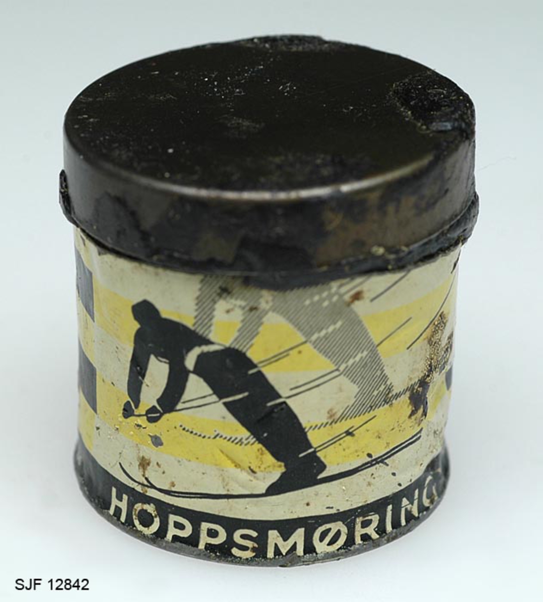 Boksen med skismurning har sylindrisk form. Den består av en metallsylinder og et lokk. Utenpå metallsylinderen er det påklistret en etikett hvor det bl. a er bilde av en skihopper som utfører et svev (øvrig informasjon fra etiketten er ført inn i feltet for påført tekst). 
Teksten som er påskrevet øverset på boksen er nesten helt borte, slik at det er umulig å tyde hva som har stått der. Etter hvert som man har brukt smurning, har det vært nødverndig å rive av deler av boksen, og dermed har deler av den påførte teksten på boksen fulgt med. 