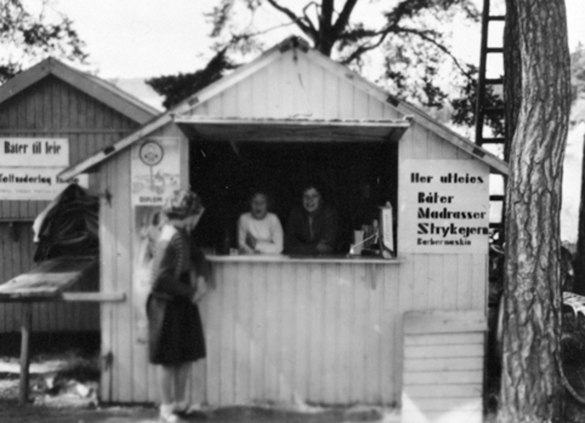 HEDMARKSMUSEET, DOMKIRKEODDEN, ROSENLUNDVIKA. KIOSK PÅ CAMPINGPLASSEN. T. V. TO UKJENTE SØATRE FRA SANDEFJORD. BEGGE PÅ BESØK TIL ONKELEN SOM DREV KIOSKEN, T. H. BERIT MOEN, PLAKAT "HER UTLEIES BÅTER, MADRASSER, STRYKEJERN, BARBERMASKIN" 1951 ELLER 1952