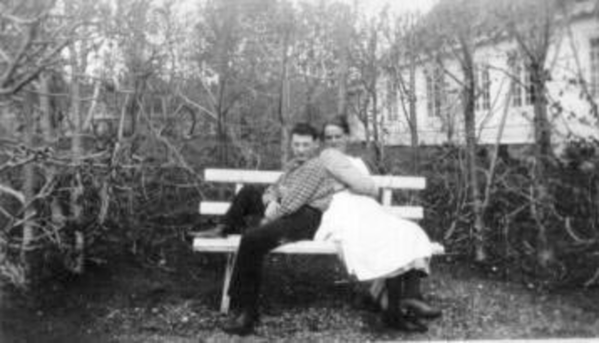 2 ungdommer på hagebenk. Hovedbygning på Eik, Helgøya bak hekken.