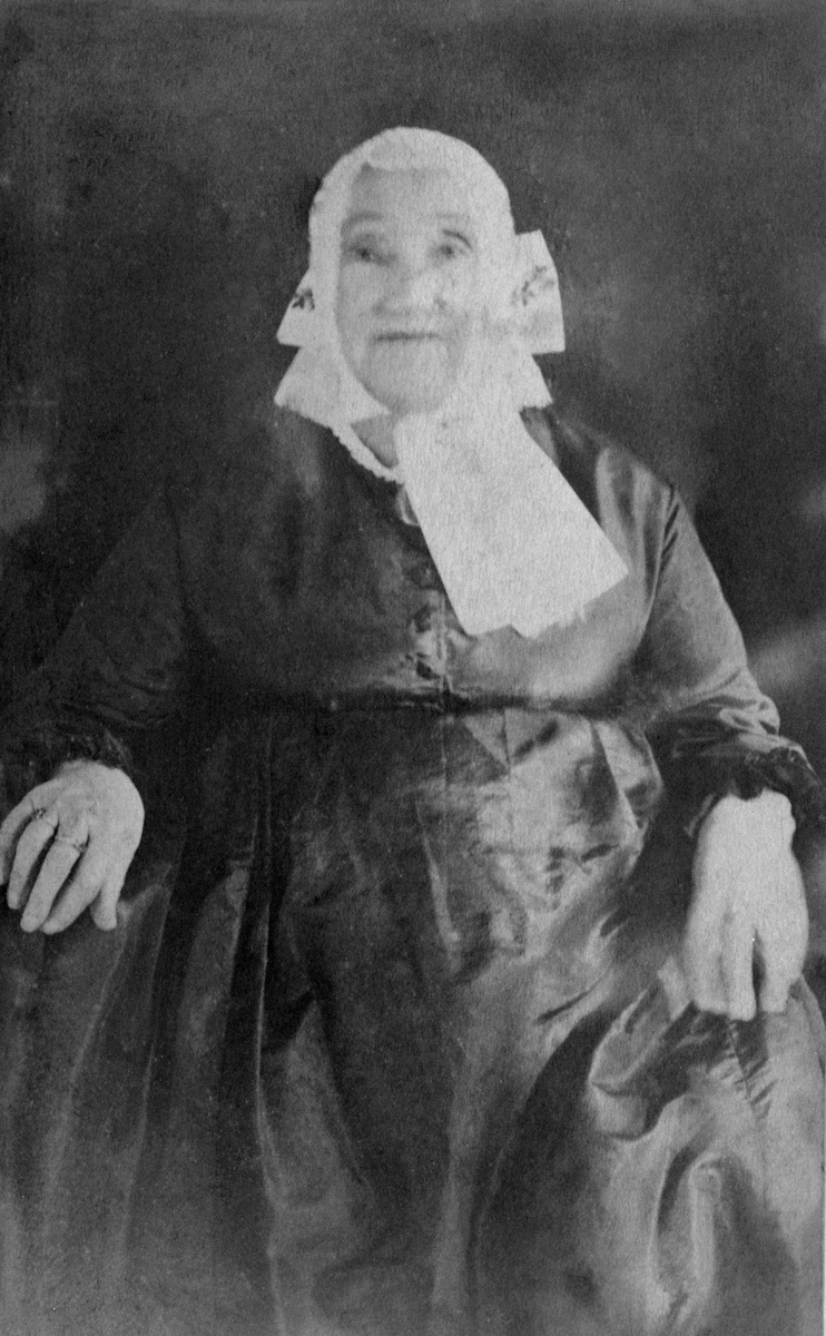 Portrett av Anne Ballishoel Kjos (født Ovren 30.9.1803- 17.02.1901). Hodeplagg, pullue med skrud, stor sløyfe i nakken og bredt band under haka, forskjøvet til høyre slik moten var fra tidligere på 1800-tallet. Mor til Øgestenius Kjos, 