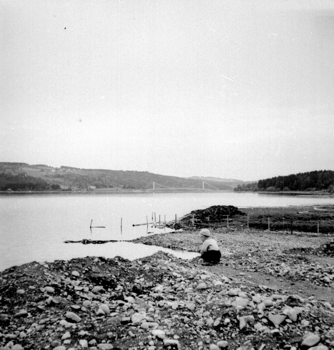 Liten gutt i Mjøskanten nedenfor strandsaga, Framnesbrua i bakgrunn, Brumunddal.