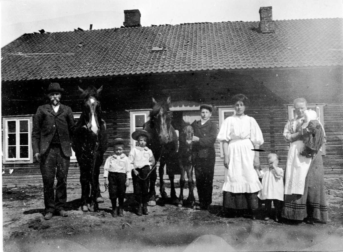 Gruppe mennesker oppstillt utenfor hus. Syverud, Ringsaker. Fra venstre er Lars Fossum f.1875, Leonard Fossum f.1906, Hans Østbye f.1905, Johan Melby f.1896, ukjent, Karen Fossum f.1909 , Gunda Fossum f.1874 med Dortha på armen. Hest med føll.