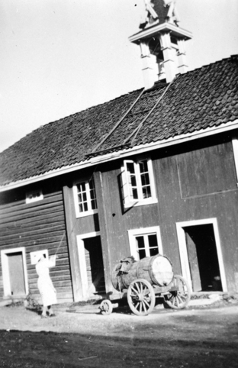 Kvinne ringer i matklokka på stabburet på Grefsheim, Nes, Hedmark. Vogn med vanntønne utenfor.