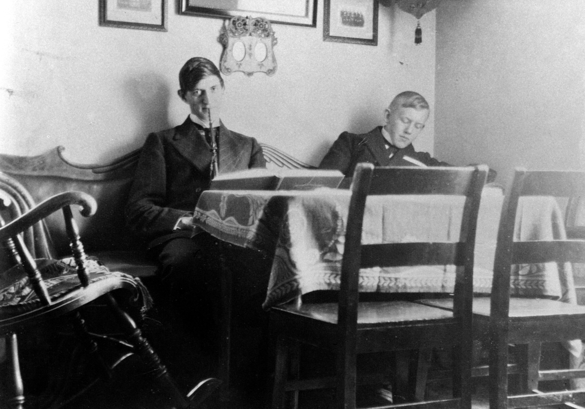 INTERIØR: STUE MED GYNGESTOL, BORD, STOLER, FOTOGRAFIER, HYLLE, GRUPPE: 2 UNGE MENN: TRYGVE FREMSTAD MED LANGPIPE, EIVIND AALSTAD, ÅLSTAD GÅRD