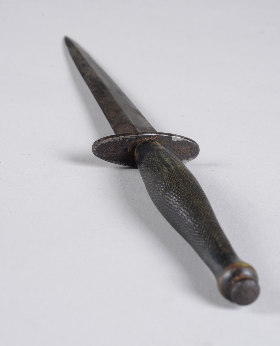 Dolk av jern i lærhylster. 
Hylsteret er forsterket i spissen og kan festes i belte. Dolken har et ruglete håndtak og et toegget blad med midtribbe.