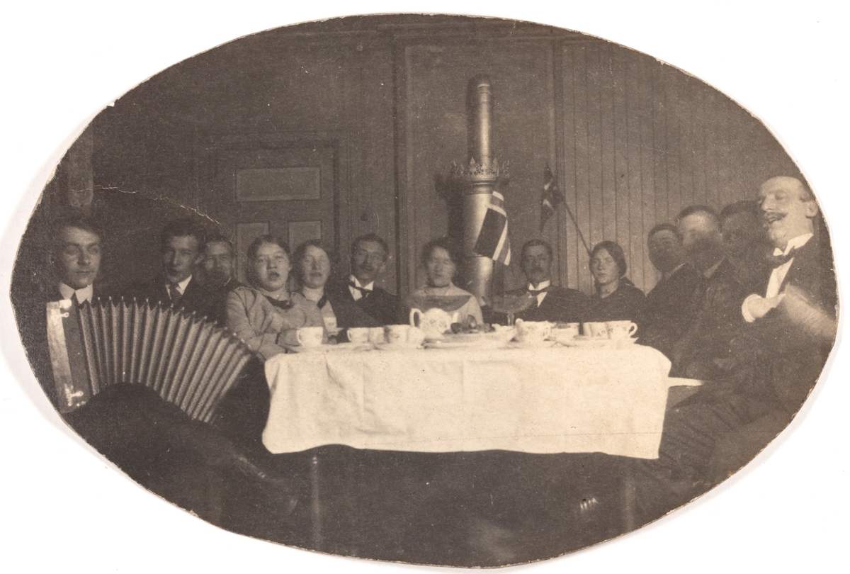 Festelig lag med mennesker rundt bord. Sang, trekkspill og flagg.
