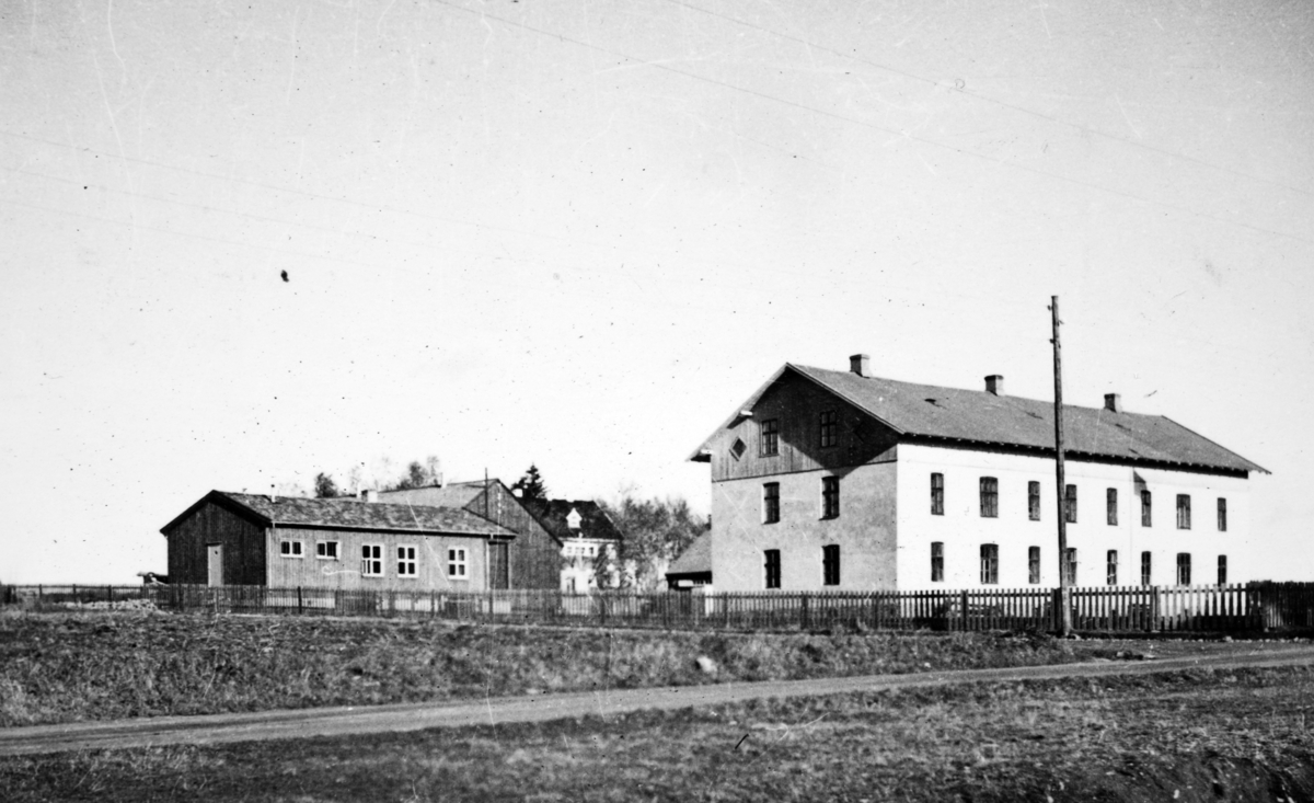 Motivet viser eksteriør av AT- leiren på Ådalsbruk, med vaktstue og kaserne i 1942. Bygningene hørte fram til 1928 til Aadals Brug Jernstøperi, og viser arbeiderboligene Murgården til høyre, ant. Tregården bakenfor, med den gamle direktørboligen, Maihaugen i bakkant. 
Se artikkel i Lautin, årbok for Løten 1993. 