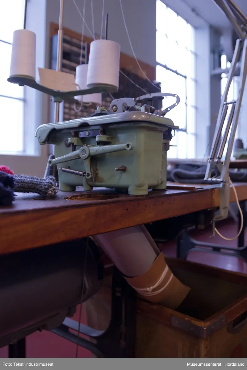 Mingrøn 1-nåls overlockmaskin som syr saman to delar trikotstoff, og samtidig kuttar av overflødig stoff med ein kniv. Maskina har tre trådar, men ein kan òg tre den med to trådar. Den har ei pumpe som oljar maskina automatisk.

Maskina er montert på eit Singer-bord saman med fleire andre symaskiner etter at den kom til museet. Bordet har eit hol der kappet frå symaskinene skulle dette ned.