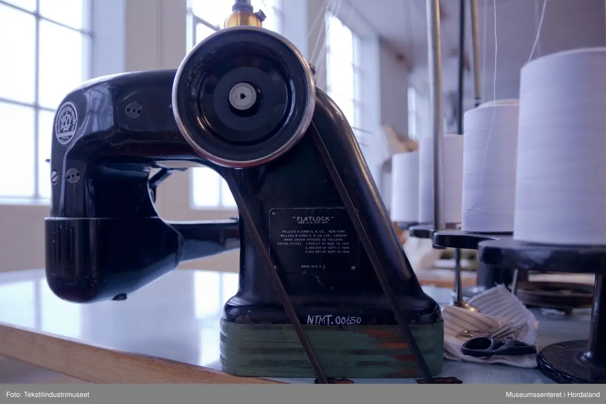 Flatlock-symaskin i svartmåla støypejern og med halvautomatisk oljing. Maskina er montert på eit Juc-bord og har Singer-motor (truleg ikkje original).

Motor: Singer alternating current electric transmitter. No. K 692357 R
Volts: 210-230.