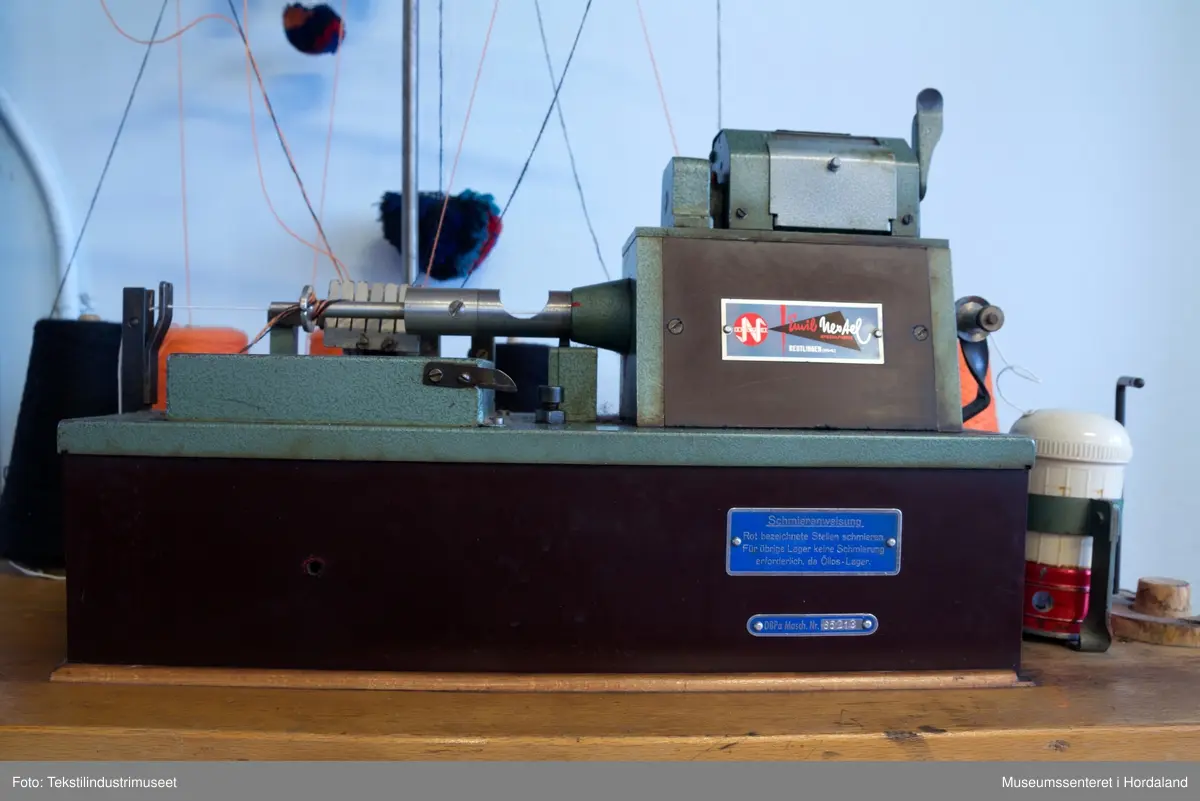 Grøn og brun maskin som lagar duskar/pompongar. Maskina har trådstativ med seks trådførarar, med ein kvit bomullstråd inni dusken og teljeapparat montert på toppen. Den står på eit Singer-bord, og av-på knappen er fastmontert i sjølve bordet.