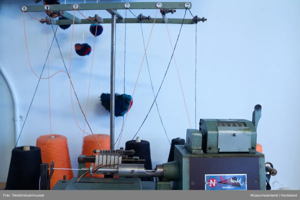 Grøn og brun maskin som lagar duskar/pompongar. Maskina har trådstativ med seks trådførarar, med ein kvit bomullstråd inni dusken og teljeapparat montert på toppen. Den står på eit Singer-bord, og av-på knappen er fastmontert i sjølve bordet.