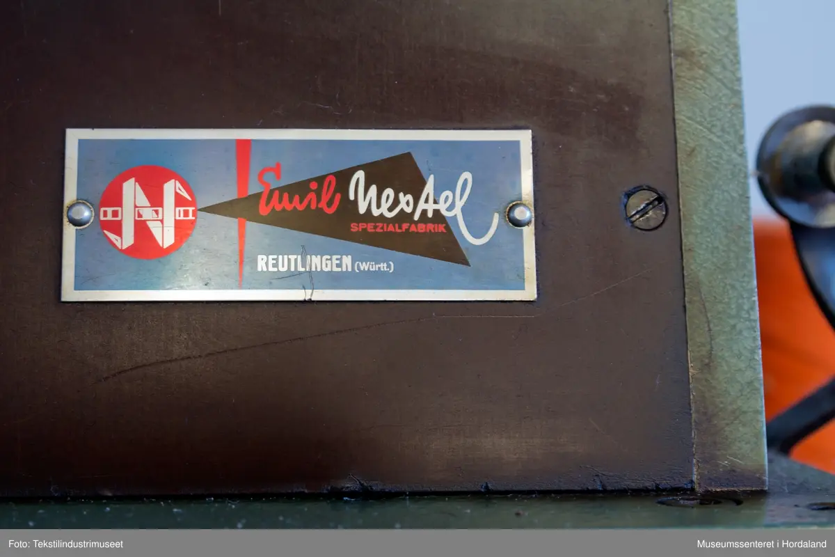 Grøn og brun maskin som lagar duskar/pompongar. Maskina har trådstativ med seks trådførarar, med ein kvit bomullstråd inni dusken og teljeapparat montert på toppen. Den står på eit Singer-bord, og av-på knappen er fastmontert i sjølve bordet.