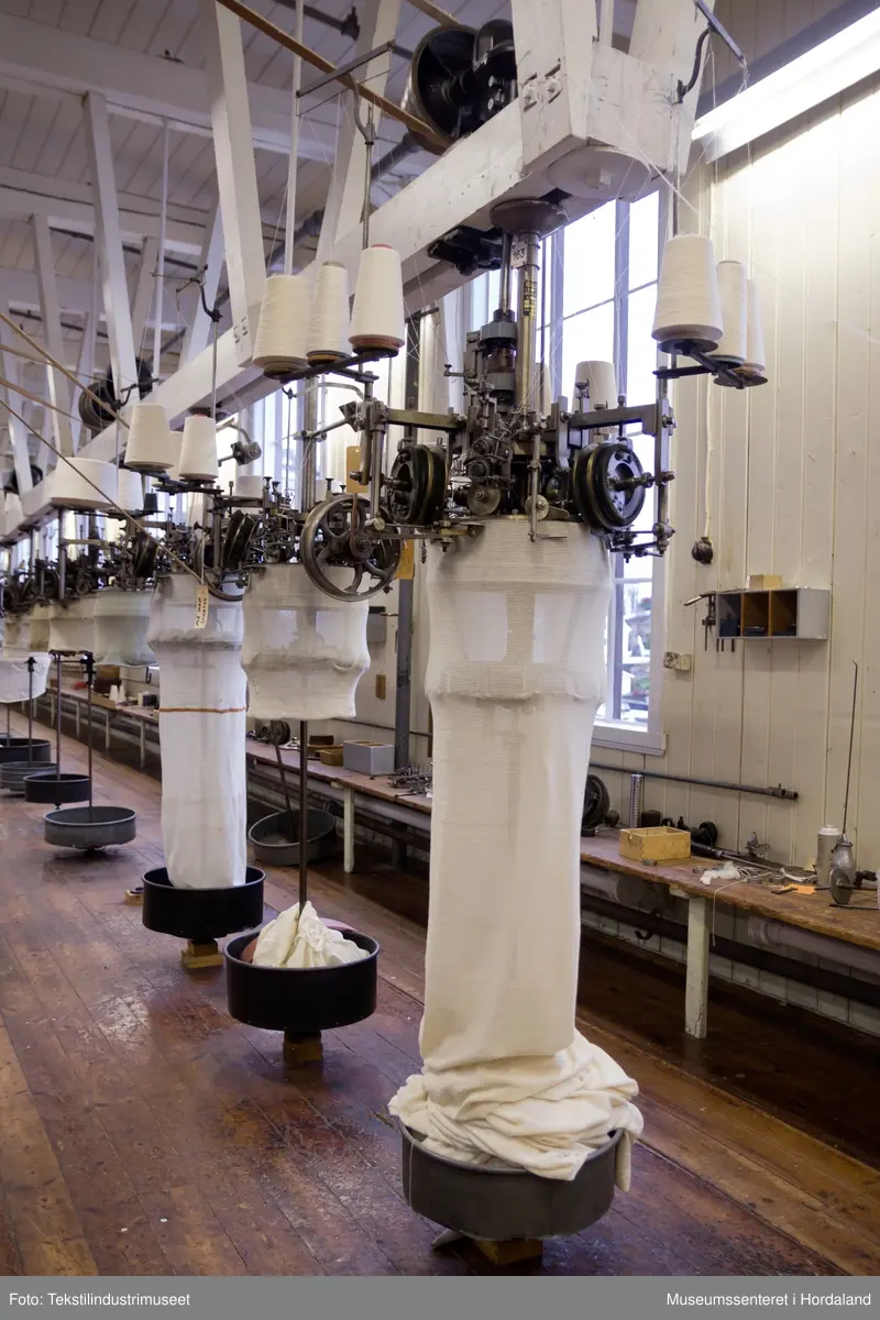 Rundstrikkemaskin/rundstol som er montert med eit feste og er kopla til reimdrift i taket. Maskina har tre system og ei nedtrekksmekanisme nederst med små metallpiggar montert på tekstilt materiale.