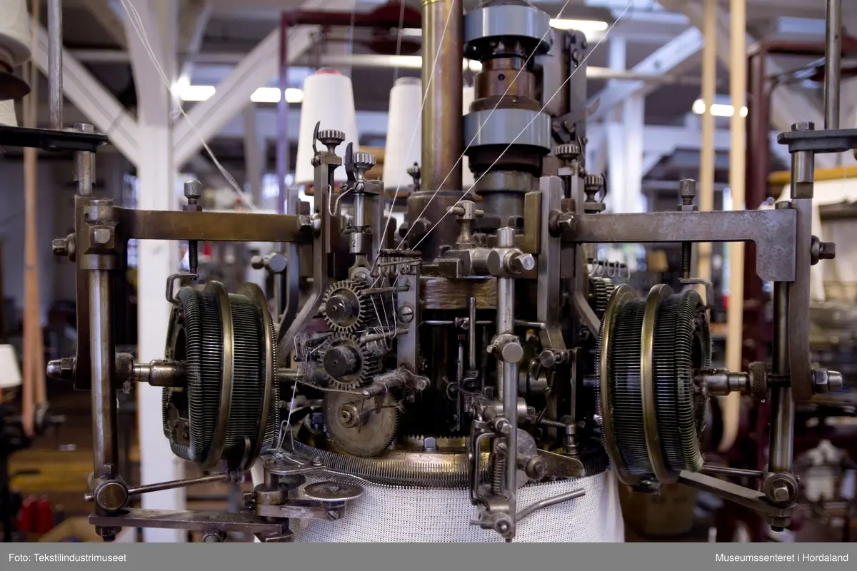 Rundstrikkemaskin/rundstol som er montert med eit feste og er kopla til reimdrift i taket. Maskina har tre system og ei nedtrekksmekanisme nederst med små metallpiggar montert på tekstilt materiale.