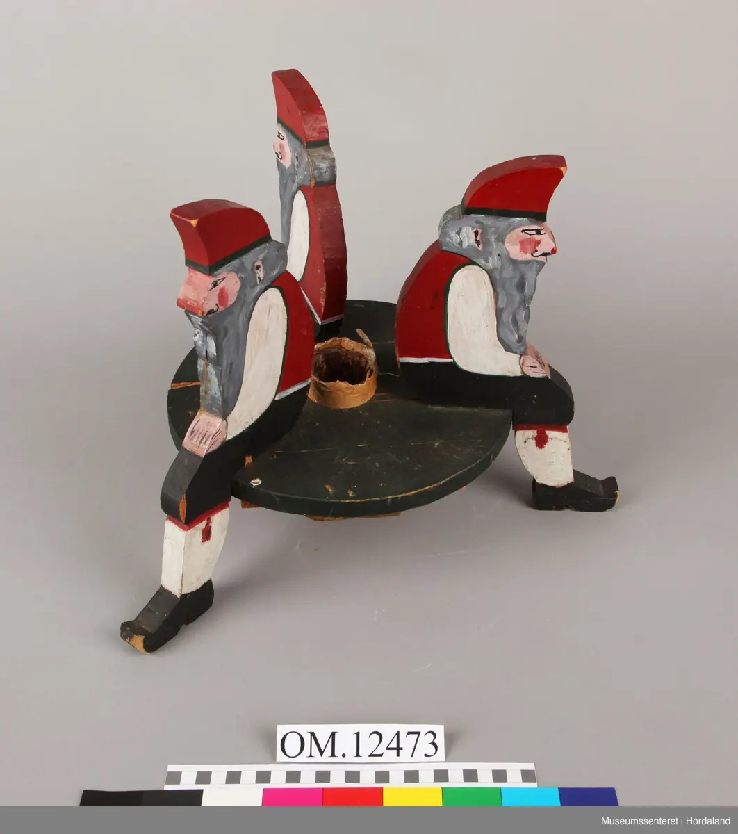 3 nisser i utskårne i tre som sit på ei rund plate. Nissaane og plata er skårne ut i 2 cm tjukk plank. Nissane er malte i ulike fargar. Plata er malt svart. Brun tape rund holet til juletreet.